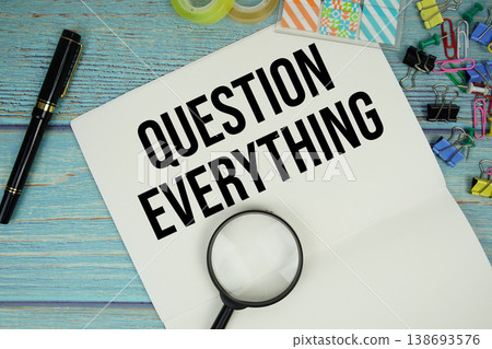 Question Everything text on a paper surrounded by office supplies and magnifying glass on a blue wooden surface 138693576