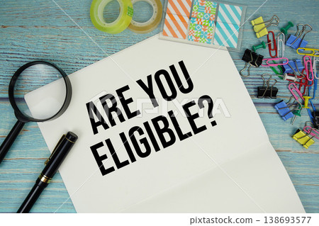 Are You Eligible? text on a paper surrounded by office supplies and magnifying glass on a blue wooden surface Are You Eligible? text on a paper surrounded by office supplies and magnifying glass on a blue wooden surface 138693577