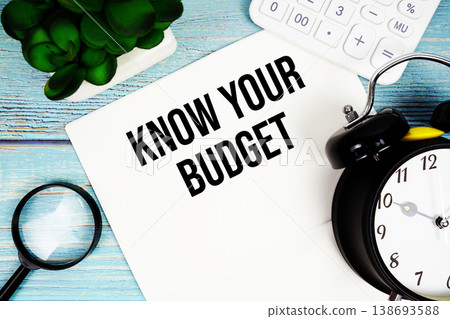 Know Your Budget written on paper placed on a blue wooden surface, alongside a magnifying glass, a clock, a calculator, and a small plant 138693588