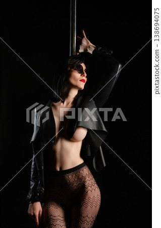 Sensual woman in open leather jacket, fishnets, and sunglasses posing with a pole on a black background. Sensual woman in open leather jacket, fishnets, and sunglasses posing with a pole on a black background. 138694075