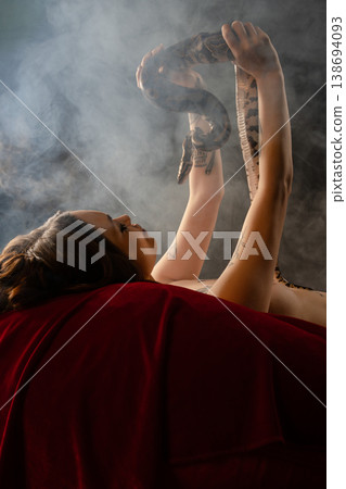 Tattooed woman lying on red velvet holding a large snake in a smoky atmosphere. Tattooed woman lying on red velvet holding a large snake in a smoky atmosphere. 138694093