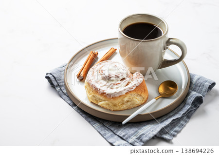 Freshly baked cinnamon roll with white icing and a cup of black coffee on a light marble background. Freshly baked cinnamon roll with white icing and a cup of black coffee on a light marble background. 138694226