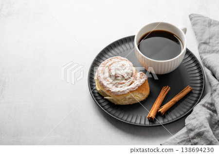Freshly baked cinnamon roll with white icing and a cup of black coffee on a light background. Freshly baked cinnamon roll with white icing and a cup of black coffee on a light background. 138694230
