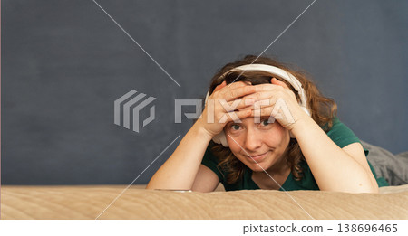 Young woman lying on bed with wireless headphones, smiling at camera while relaxed and enjoying music at home in a cozy, casual moment Young woman lying on bed with wireless headphones, smiling at camera while relaxed and enjoying music at home in a cozy, casual moment 138696465