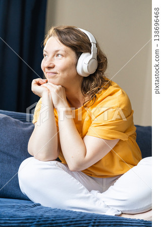 Woman in a yellow shirt and white pants sitting comfortably on a sofa, smiling while listening to music with headphones. Young happy woman enjoying music Woman in a yellow shirt and white pants sitting comfortably on a sofa, smiling while listening to music with headphones. Young happy woman enjoying music 138696468