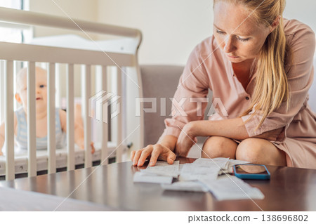 Mother sitting on sofa calculating monthly expenses with receipts while baby lies in crib nearby at home. Stressed mom managing budget and childcare. Financial pressure and parenthood concept 138696802