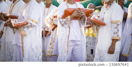 Participants in white clothing line up holding folded orange robes as soft sunlight reveals intricate lace details and hands within a medium to large ceremonial setting. Participants in white clothing line up holding folded orange robes as soft sunlight reveals intricate lace details and hands within a medium to large ceremonial setting. 138697641