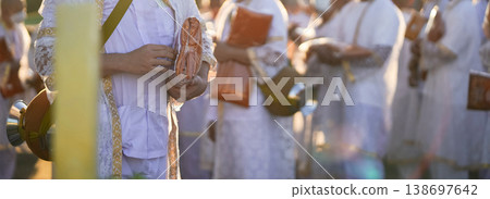 Participants in white clothing line up holding folded orange robes as soft sunlight reveals intricate lace details and hands within a medium to large ceremonial setting. Participants in white clothing line up holding folded orange robes as soft sunlight reveals intricate lace details and hands within a medium to large ceremonial setting. 138697642