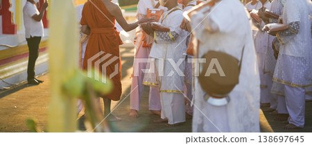 Participants in white clothing line up holding folded orange robes as soft sunlight reveals intricate lace details and hands within a medium to large ceremonial setting. 138697645