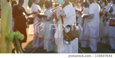 Participants in white clothing line up holding folded orange robes as soft sunlight reveals intricate lace details and hands within a medium to large ceremonial setting. 138697648