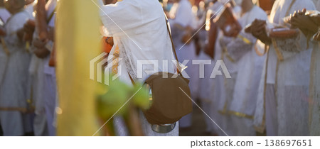 Participants in white clothing line up holding folded orange robes as soft sunlight reveals intricate lace details and hands within a medium to large ceremonial setting. 138697651