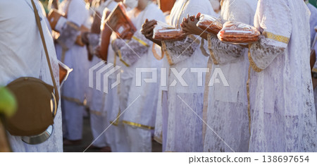 Participants in white clothing line up holding folded orange robes as soft sunlight reveals intricate lace details and hands within a medium to large ceremonial setting. Participants in white clothing line up holding folded orange robes as soft sunlight reveals intricate lace details and hands within a medium to large ceremonial setting. 138697654