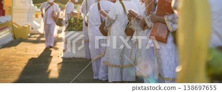Participants in white clothing line up holding folded orange robes as soft sunlight reveals intricate lace details and hands within a medium to large ceremonial setting. Participants in white clothing line up holding folded orange robes as soft sunlight reveals intricate lace details and hands within a medium to large ceremonial setting. 138697655