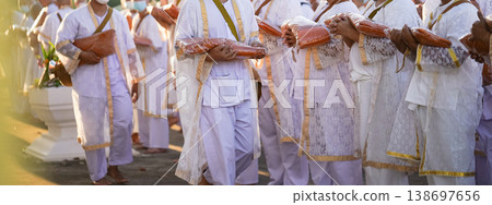 Participants in white clothing line up holding folded orange robes as soft sunlight reveals intricate lace details and hands within a medium to large ceremonial setting. Participants in white clothing line up holding folded orange robes as soft sunlight reveals intricate lace details and hands within a medium to large ceremonial setting. 138697656