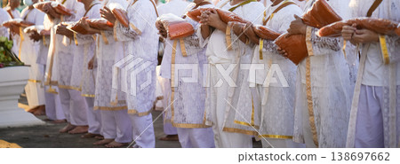 Participants in white clothing line up holding folded orange robes as soft sunlight reveals intricate lace details and hands within a medium to large ceremonial setting. Participants in white clothing line up holding folded orange robes as soft sunlight reveals intricate lace details and hands within a medium to large ceremonial setting. 138697662