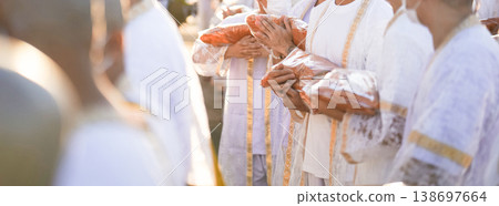 Participants in white clothing line up holding folded orange robes as soft sunlight reveals intricate lace details and hands within a medium to large ceremonial setting. Participants in white clothing line up holding folded orange robes as soft sunlight reveals intricate lace details and hands within a medium to large ceremonial setting. 138697664