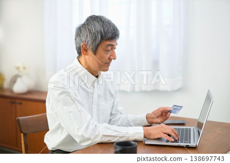 一位老年男子一邊看著自己的信用卡,一邊在電腦上進行信用卡付款。 一位老年男子一邊看著自己的信用卡,一邊在電腦上進行信用卡付款。 138697743