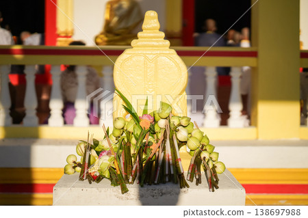 A temple marker stands above arranged lotus incense and candles as fresh lotus bundles and folded robes rest nearby with a blurred statue adding depth to the warm sacred ceremonial setting. 138697988