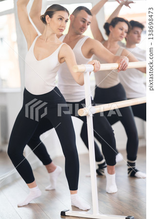 Group of sports people standing in row together while doing ballet dance moves demi plie while holding on ballet barre in ballet studio 138698473