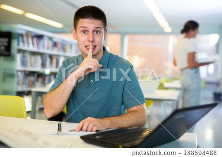 Young casual man placing fingers on lips with shhh while using computer in the library Young casual man placing fingers on lips with shhh while using computer in the library 138698488