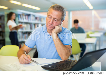 Tired man in casual clothes working remotely using a computer and in a quiet library Tired man in casual clothes working remotely using a computer and in a quiet library 138698500