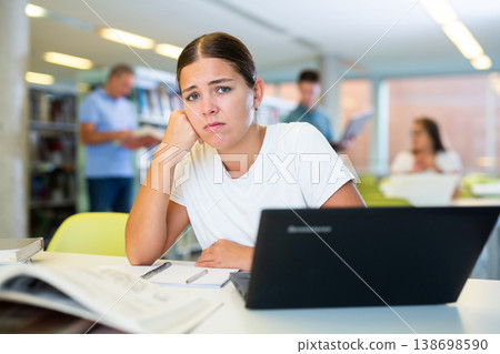 Tired casual female remote worker using laptop and reading books in the library Tired casual female remote worker using laptop and reading books in the library 138698590