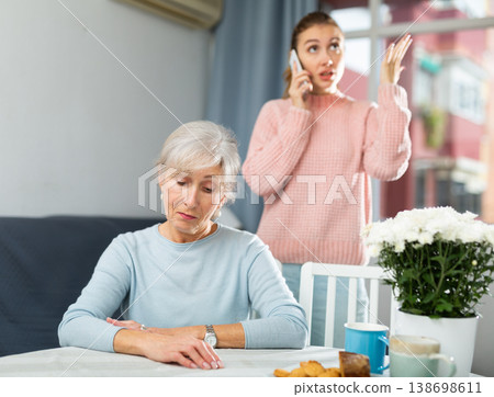 Upset senior woman sitting at table while irritated adult daughter talking on phone Upset senior woman sitting at table while irritated adult daughter talking on phone 138698611