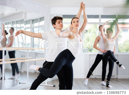 Pretty female ballerina and male partner practicing ballet arabesque pose during training with group active people in dancing hall 138698773