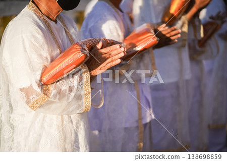 Thai men in white walk in line with palms together carrying folded orange monk robes as medium to large group approaches temple doorway, warm light before ordination. Thai men in white walk in line with palms together carrying folded orange monk robes as medium to large group approaches temple doorway, warm light before ordination. 138698859