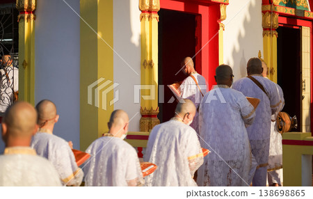 Thai men in white walk in line with palms together carrying folded orange monk robes as medium to large group approaches temple doorway, warm light before ordination. Thai men in white walk in line with palms together carrying folded orange monk robes as medium to large group approaches temple doorway, warm light before ordination. 138698865