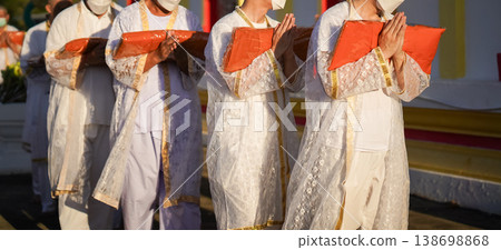 Thai men in white walk in line with palms together carrying folded orange monk robes as medium to large group approaches temple doorway, warm light before ordination. Thai men in white walk in line with palms together carrying folded orange monk robes as medium to large group approaches temple doorway, warm light before ordination. 138698868