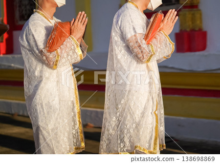 Thai men in white walk in line with palms together carrying folded orange monk robes as medium to large group approaches temple doorway, warm light before ordination. Thai men in white walk in line with palms together carrying folded orange monk robes as medium to large group approaches temple doorway, warm light before ordination. 138698869