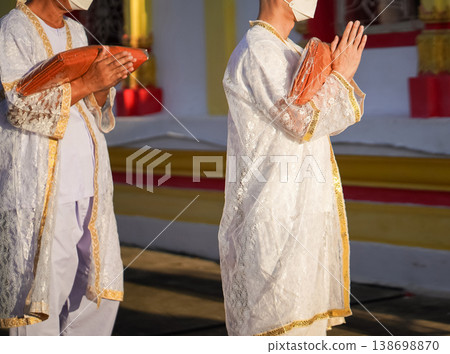 Thai men in white walk in line with palms together carrying folded orange monk robes as medium to large group approaches temple doorway, warm light before ordination. Thai men in white walk in line with palms together carrying folded orange monk robes as medium to large group approaches temple doorway, warm light before ordination. 138698870