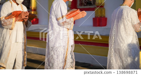 Thai men in white walk in line with palms together carrying folded orange monk robes as medium to large group approaches temple doorway, warm light before ordination. Thai men in white walk in line with palms together carrying folded orange monk robes as medium to large group approaches temple doorway, warm light before ordination. 138698871