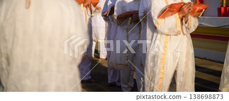 Thai men in white walk in line with palms together carrying folded orange monk robes as medium to large group approaches temple doorway, warm light before ordination. 138698873
