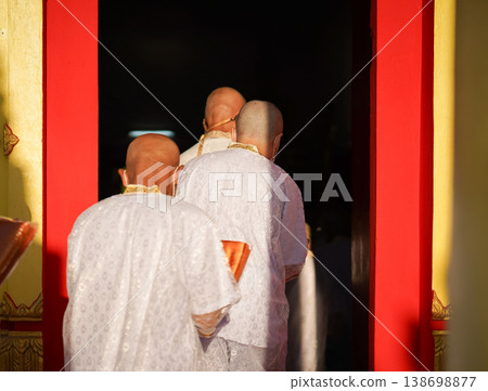 Thai men in white walk in line with palms together carrying folded orange monk robes as medium to large group approaches temple doorway, warm light before ordination. Thai men in white walk in line with palms together carrying folded orange monk robes as medium to large group approaches temple doorway, warm light before ordination. 138698877