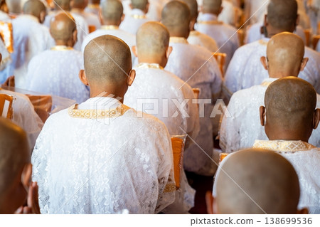 A rear perspective shows white clad ordinands seated in rows inside the temple holding orange robes with shaved heads and folded hands in a soft blurred ritual setting. 138699536