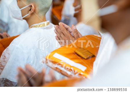 A rear perspective shows white clad ordinands seated in rows inside the temple holding orange robes with shaved heads and folded hands in a soft blurred ritual setting. 138699538