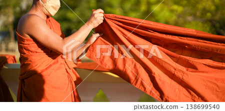 A senior monk adjusts a novice robe as hands tighten the woven belt around orange cloth in sunlight with focus on texture folds and precise fitting. A senior monk adjusts a novice robe as hands tighten the woven belt around orange cloth in sunlight with focus on texture folds and precise fitting. 138699540