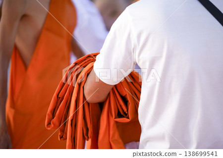 A senior monk adjusts a novice robe as hands tighten the woven belt around orange cloth in sunlight with focus on texture folds and precise fitting. 138699541