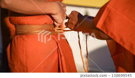 A senior monk adjusts a novice robe as hands tighten the woven belt around orange cloth in sunlight with focus on texture folds and precise fitting. A senior monk adjusts a novice robe as hands tighten the woven belt around orange cloth in sunlight with focus on texture folds and precise fitting. 138699545