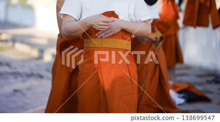 A senior monk adjusts a novice robe as hands tighten the woven belt around orange cloth in sunlight with focus on texture folds and precise fitting. 138699547