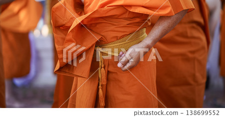 A senior monk adjusts a novice robe as hands tighten the woven belt around orange cloth in sunlight with focus on texture folds and precise fitting. 138699552