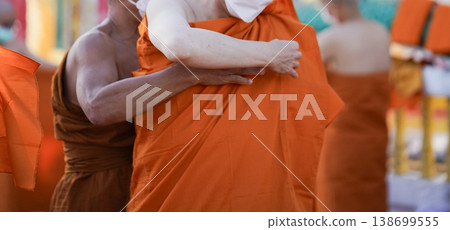 A senior monk adjusts a novice robe as hands tighten the woven belt around orange cloth in sunlight with focus on texture folds and precise fitting. 138699555