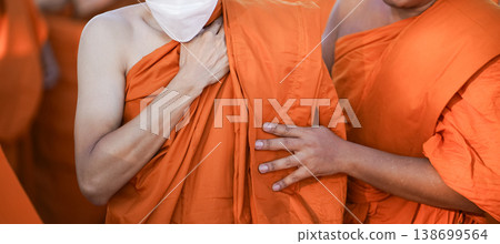 A senior monk adjusts a novice robe as hands tighten the woven belt around orange cloth in sunlight with focus on texture folds and precise fitting. 138699564