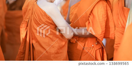 A senior monk adjusts a novice robe as hands tighten the woven belt around orange cloth in sunlight with focus on texture folds and precise fitting. A senior monk adjusts a novice robe as hands tighten the woven belt around orange cloth in sunlight with focus on texture folds and precise fitting. 138699565