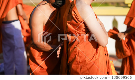 A senior monk adjusts a novice robe as hands tighten the woven belt around orange cloth in sunlight with focus on texture folds and precise fitting. A senior monk adjusts a novice robe as hands tighten the woven belt around orange cloth in sunlight with focus on texture folds and precise fitting. 138699566