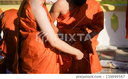 A senior monk adjusts a novice robe as hands tighten the woven belt around orange cloth in sunlight with focus on texture folds and precise fitting. A senior monk adjusts a novice robe as hands tighten the woven belt around orange cloth in sunlight with focus on texture folds and precise fitting. 138699567