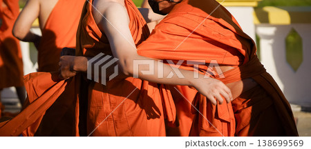 A senior monk adjusts a novice robe as hands tighten the woven belt around orange cloth in sunlight with focus on texture folds and precise fitting. 138699569