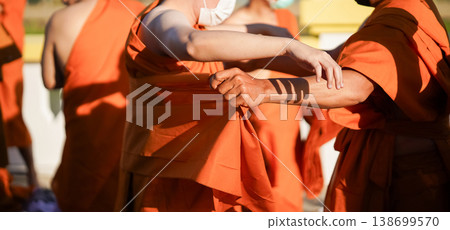 A senior monk adjusts a novice robe as hands tighten the woven belt around orange cloth in sunlight with focus on texture folds and precise fitting. A senior monk adjusts a novice robe as hands tighten the woven belt around orange cloth in sunlight with focus on texture folds and precise fitting. 138699570